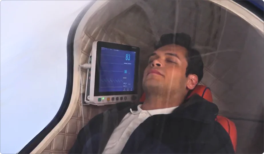 A man reclines inside the Apex hyperbaric oxygen therapy (HBOT) chamber with his eyes closed. A monitor next to him displays his vital signs, including a heart rate of 83. The chamber appears to be sealed and padded for comfort.