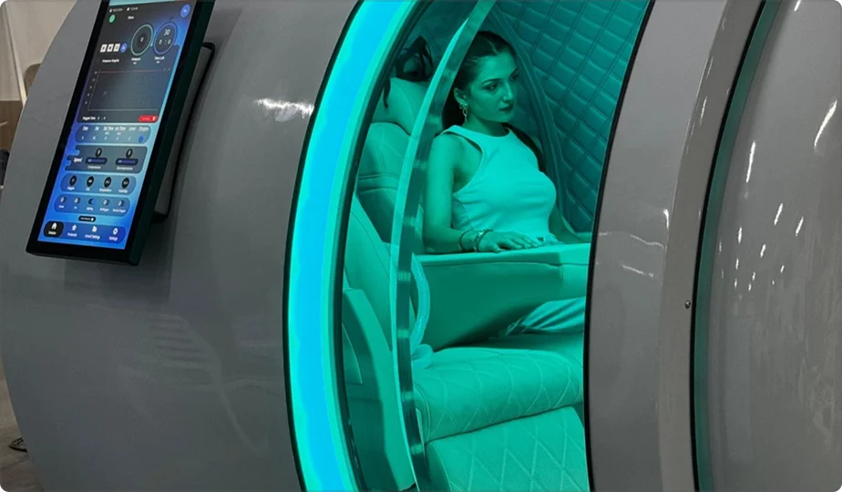A woman in a white dress sits inside the Atlantis hyperbaric oxygen therapy (HBOT) chamber with teal lighting, surrounded by padded walls and a touchscreen control panel mounted outside.