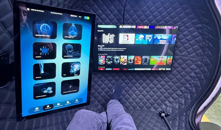 A person sits in a padded, pod-like chair with their legs visible. In front of them are two screens: a large vertical touchscreen displaying scientific images, and a horizontal TV displaying movies and games.