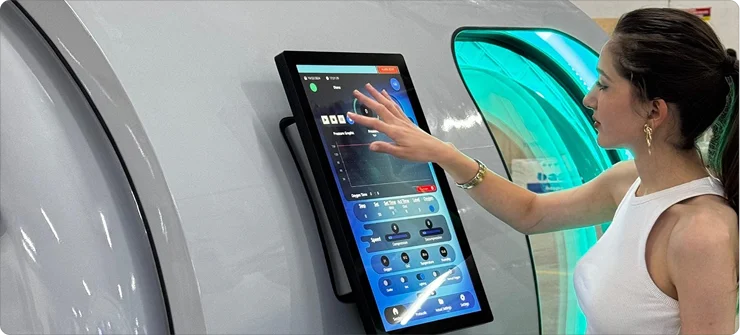 A woman in a white sleeveless top interacts with a large touchscreen control panel on the Atlantis hyperbaric oxygen therapy (HBOT) chamber.
