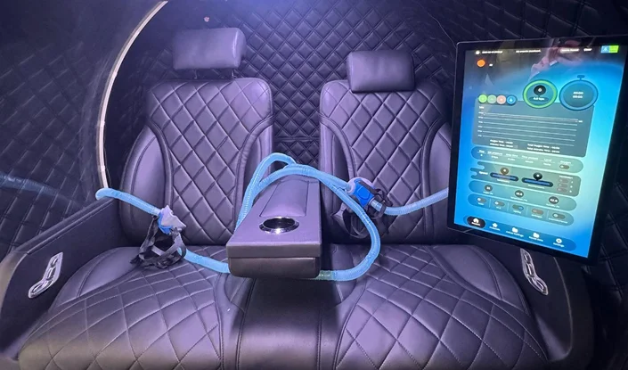 Two black, diamond-stitched leather seats inside the Atlantis hyperbaric oxygen therapy (HBOT) chamber, separated by an armrest with cup holder. Blue seatbelts are draped across, and a large touchscreen display with controls is mounted on the right.