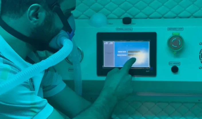 A person wearing a breathing mask touches a touchscreen control panel inside the Atlantis hyperbaric oxygen therapy (HBOT) chamber. The lighting has a blue-green tint.