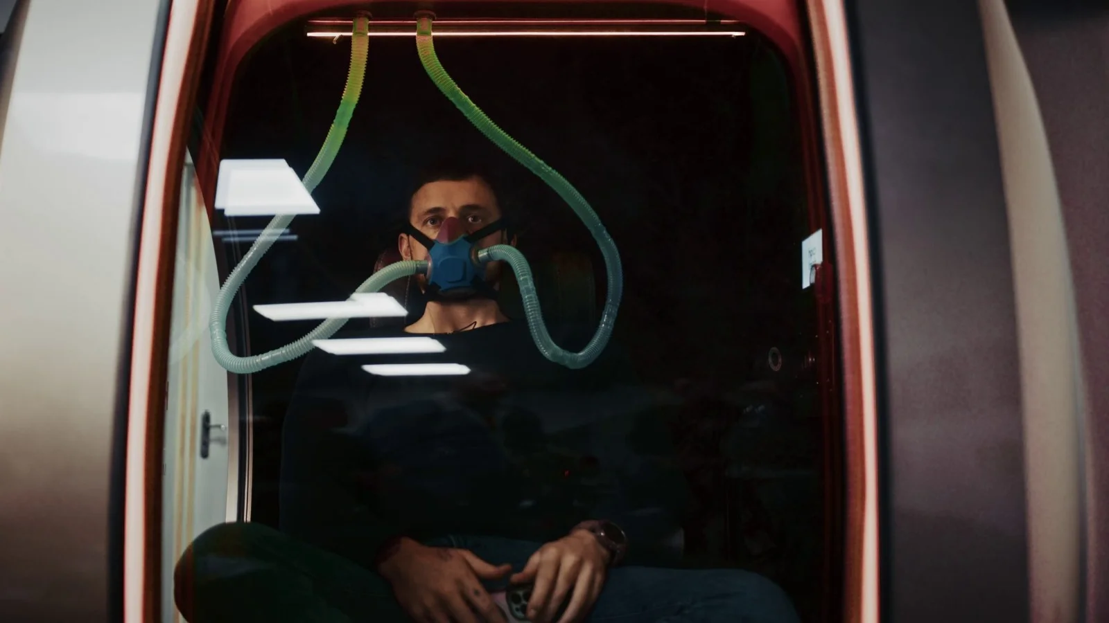 Vegan filmmaker Jordan Mulligan sits inside a hyperbaric oxygen chamber wearing a blue mask with green tubes attached to it, looking forward. The image is taken through a window, reflecting some lights on the glass.