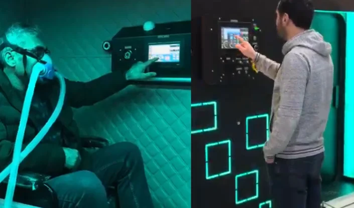 Two men interact with the interior and exterior controls of the Matrix hyperbaric oxygen therapy (HBOT) machine. On the left, a man sits wearing a breathing mask and adjusts a touchscreen. On the right, another man stands and touches a different brightly lit control panel.