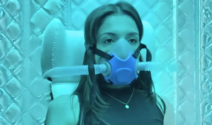 A woman wearing a blue oxygen mask with tubes sits against a padded, metallic-looking inside the Matrix hyperbaric oxygen therapy (HBOT) chamber, illuminated by blue lighting.