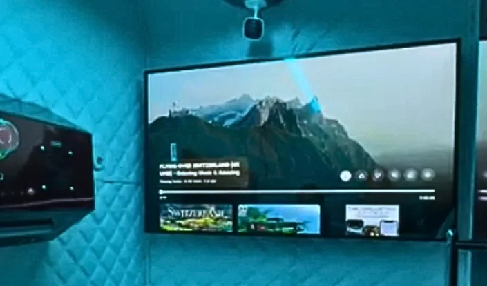 The interiors of the Matrix hyperbaric oxygen therapy (HBOT) chamber with blue lighting features a large flat-screen TV mounted on the wall, displaying a mountain landscape. A projector is mounted above the TV, and there is an air conditioner to the left.