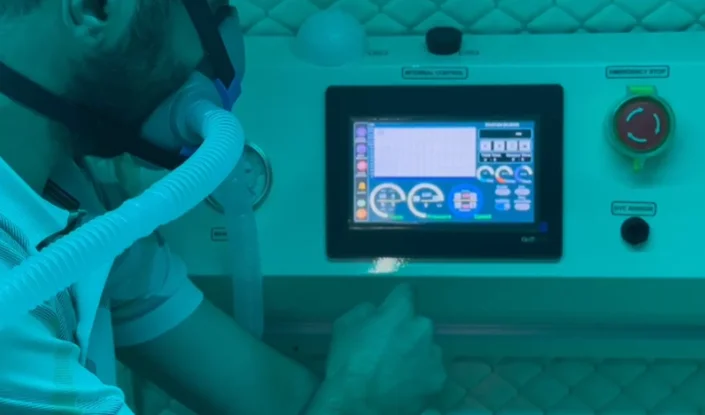 A person wearing a breathing mask adjusts controls on a digital touchscreen panel with various gauges and buttons in the blue-lit environment of the Matrix hyperbaric oxygen therapy (HBOT) chamber.