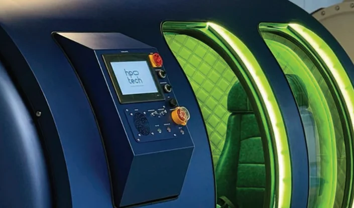 A close-up of the Mediterranean hyperbaric oxygen therapy (HBOT) chamber with a digital control panel and a green-lit interior featuring a padded chair.