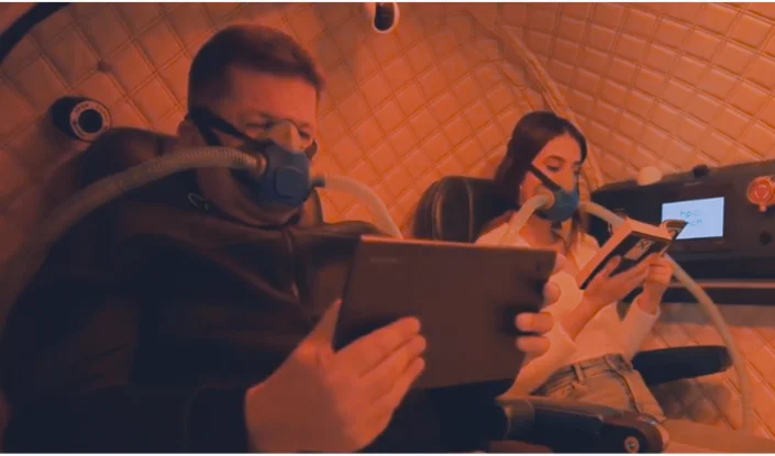 Two people sit in reclining chairs inside the Mediterranean hyperbaric oxygen therapy (HBOT) chamber, wearing masks with attached tubes. The man reads from a tablet while the woman reads a book. The environment is dimly lit with red lighting.