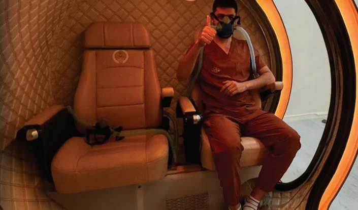 A person wearing a mask and scrubs sits inside the Mediterranean hyperbaric oxygen therapy (HBOT) chamber, giving a thumbs-up gesture. An empty chair is beside them, and soft orange lighting highlights the interior.