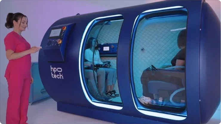 A woman in pink scrubs stands beside a large blue the Mediterranean hyperbaric oxygen therapy (HBOT) chamber with two people seated inside, both wearing masks, in a brightly lit, modern setting.