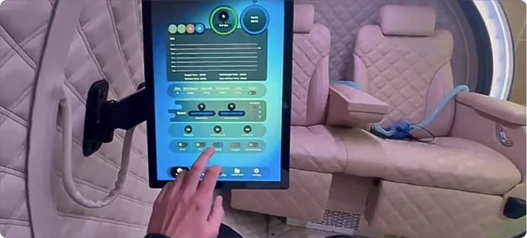 A hand interacts with a touchscreen control panel inside the luxurious, beige, quilted interior of the Mediterranean hyperbaric oxygen therapy (HBOT) chamber, featuring two plush seats and a large armrest.