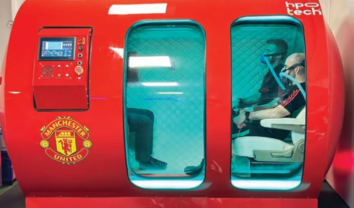 Two men are seated inside a red Mediterranean hyperbaric oxygen therapy (HBOT) chamber with a Manchester United logo, blue-lit interior, and control panel on the front.