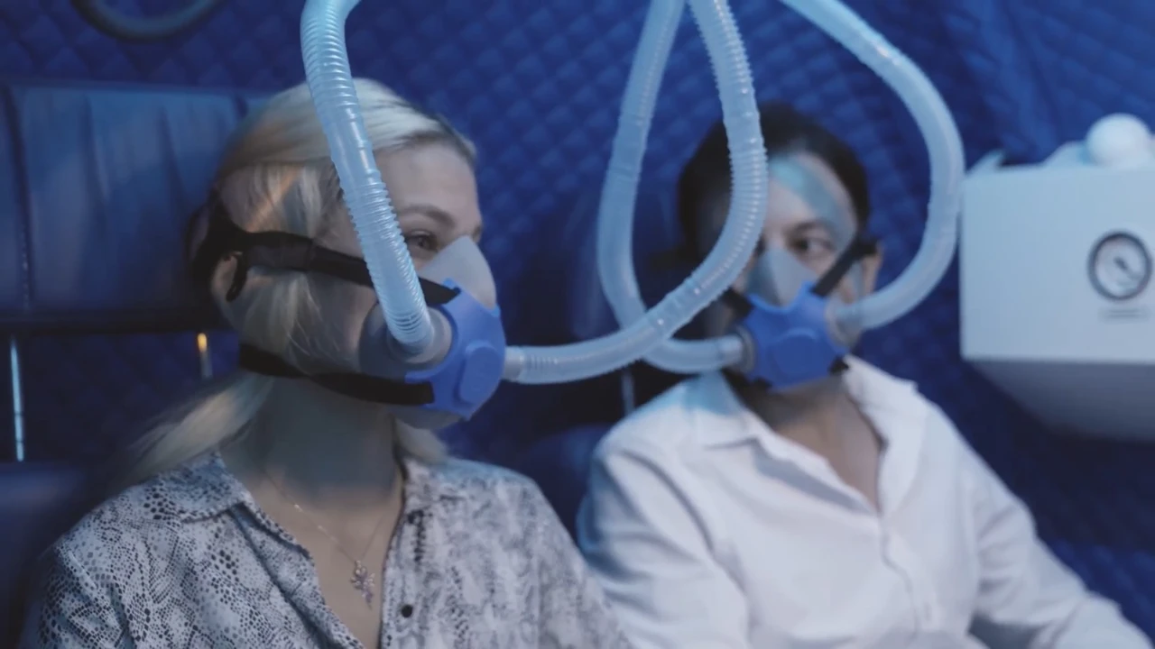 Two people sit side by side in a blue hyperbaric oxygen therapy (HBOT) chamber wearing respiratory masks connected to tubes. They appear to be in a specialized setting, possibly for oxygen therapy, and are looking at each other.