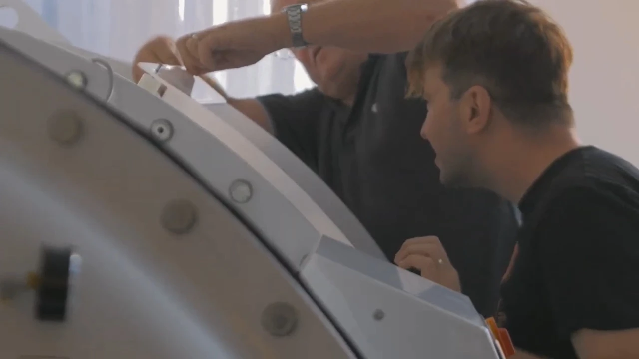 Two people work together closely, inspecting and assembling a large hyperbaric oxygen therapy machine. One person is partially visible, while the other leans in, focusing on the equipment.