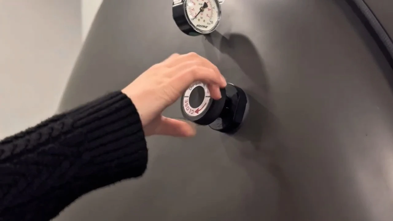 A person wearing a black sweater is turning the dial of a hyperbaric oxygen chamber. A round gauge or dial is visible above the lock on the gray metal surface.