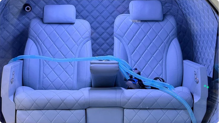 Two light blue, leather, quilted seats with headrests and armrests in the Atlantis hyperbaric oxygen therapy (HBOT) chamber, separated by a center console. A blue tube and a black device rest across the seats. The background is quilted to match the seats.