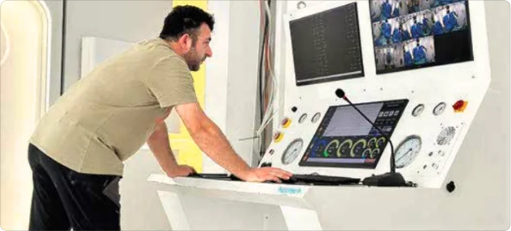 A man stands and leans over a control panel with multiple screens, gauges, and buttons, monitoring various data. Several small video feeds are displayed on the upper right screen.