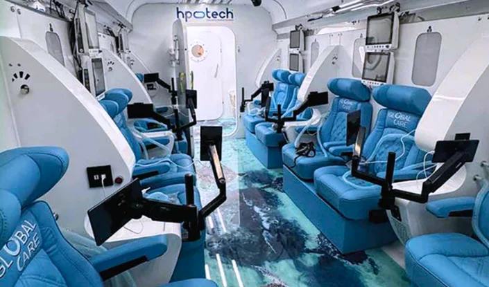 The interior of the Titan hyperbaric oxygen therapy (HBOT) chamber with blue reclining seats, individual monitors, and arm rests. The floor has an ocean-themed design and white walls with medical equipment attached.