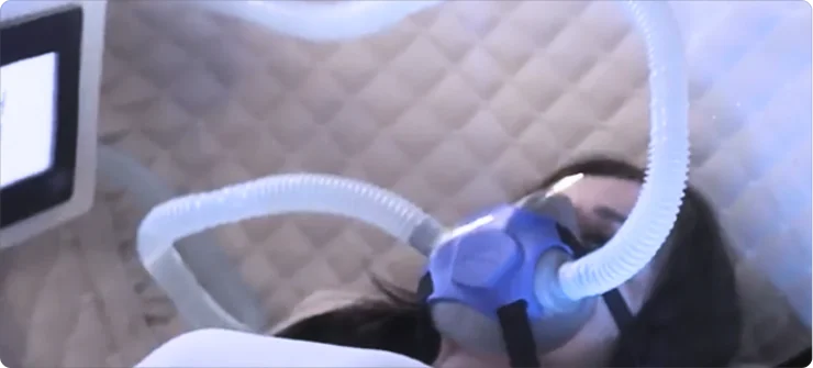 A person lies down wearing a blue oxygen mask connected to tubes, inside the padded enclosure of the Trojan hyperbaric oxygen therapy (HBOT) chamber, with a monitor visible on the side.