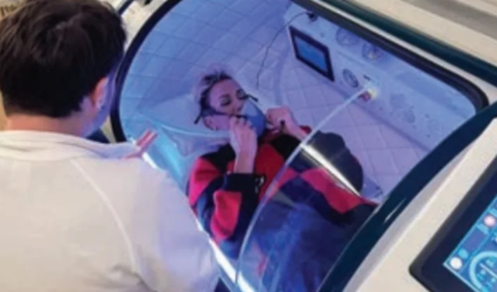 A person lies inside a Trojan hyperbaric oxygen therapy (HBOT) chamber, wearing a mask. Another person, seen from behind, appears to be assisting or speaking to them. The chamber is lit with a blue glow and medical equipment is visible.