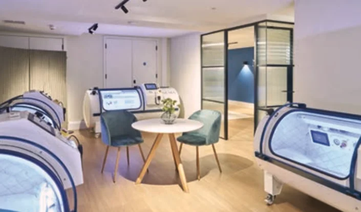 A modern medical room with several Trojan hyperbaric oxygen therapy (HBOT) chambers, a small round table, two teal chairs, and bright, clean decor.