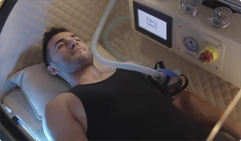 A man in a black tank top lies inside a Trojan hyperbaric oxygen therapy (HBOT) chamber with a breathing mask connected to a tube; control panels and a screen are visible next to him.