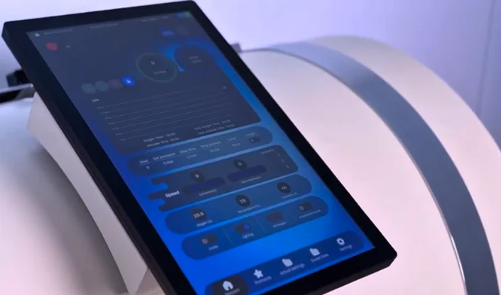 A tablet for the Trojan hyperbaric oxygen therapy (HBOT) chamber. It has a blue interface displaying various controls and settings, and rests on a white, curved surface under soft lighting.