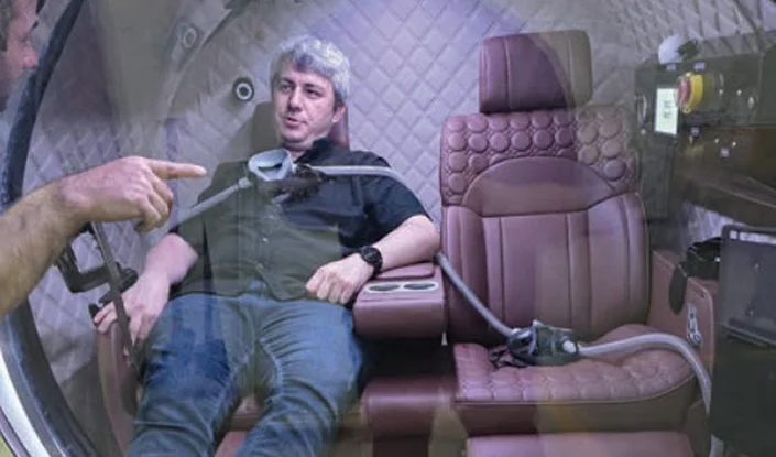 A man sits in a padded chair inside the Atlantis hyperbaric oxygen therapy (HBOT) chamber, with tubes and controls attached. Another person’s hand is visible, pointing at the man or equipment.