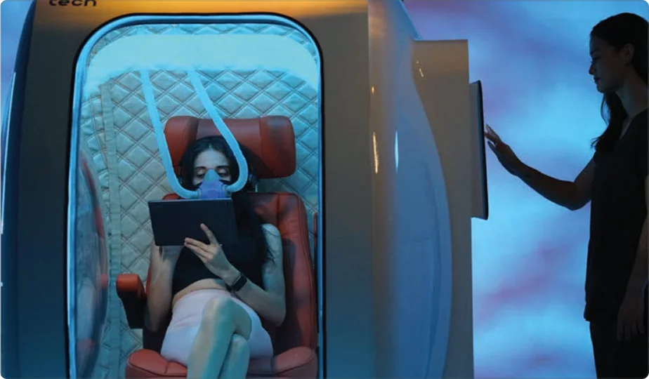 A woman sits inside the Zeugma hyperbaric oxygen therapy (HBOT) chamber, wearing an oxygen mask and holding a tablet. Another person stands outside the chamber, interacting with a touchscreen panel. The scene is lit with a blue ambient glow.