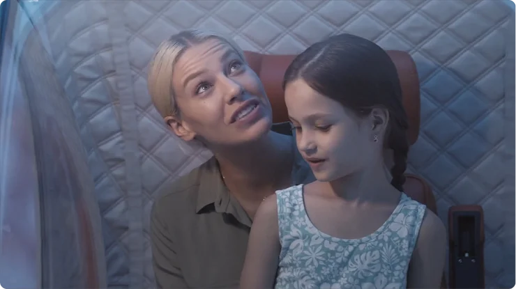 A woman with blonde hair and a young girl with brown hair sit together inside the Zeugma hyperbaric oxygen therapy (HBOT) chamber. The woman looks up and smiles, while the girl, wearing a patterned dress, sits on her lap and looks to the side.