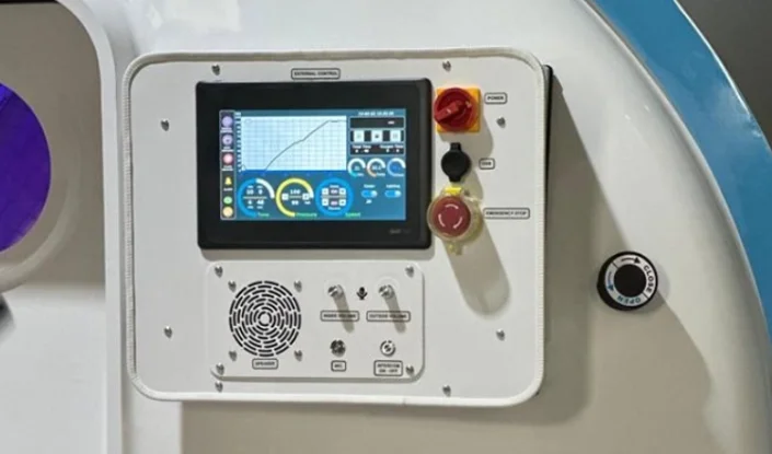 A close-up of a white machine control panel with a touchscreen displaying charts and controls, several buttons and switches, and a speaker grille below the display.