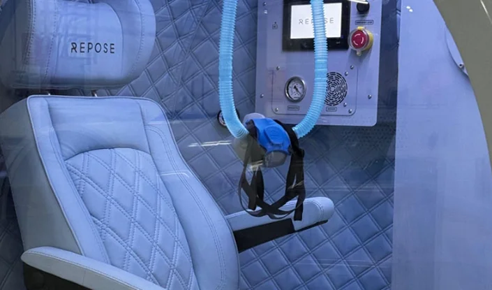 A padded light blue chair sits inside the Zeugma hyperbaric oxygen therapy (HBOT) chamber. There’s a breathing mask attached to blue hoses hanging in front of the chair, and a control panel with dials and buttons is mounted nearby.