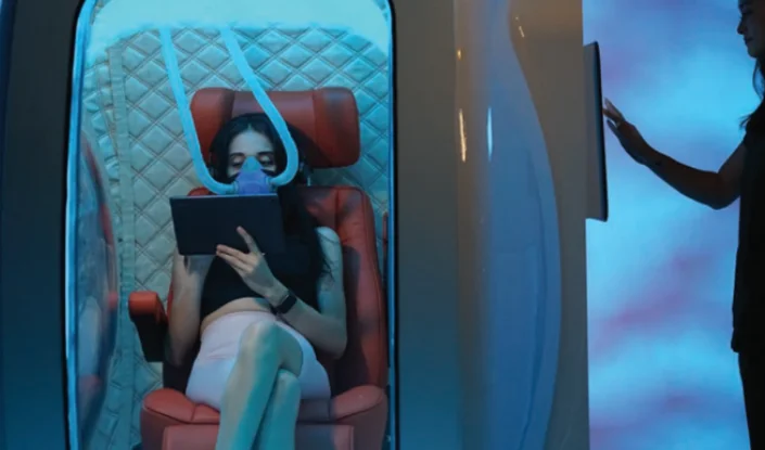 A woman sits in the Zeugma hyperbaric oxygen therapy (HBOT) chamber, wearing a breathing mask and reading a tablet. Another person stands outside, interacting with a touchscreen on the pod. The scene is lit with blue ambient lighting.