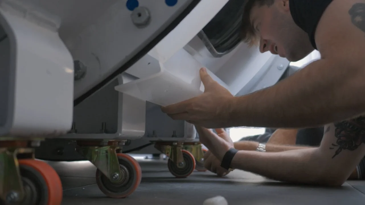 A person lies on the floor, working underneath a hyperbaric oxygen therapy (HBOT) chamber. He is holding a white plastic tray and making adjustments.